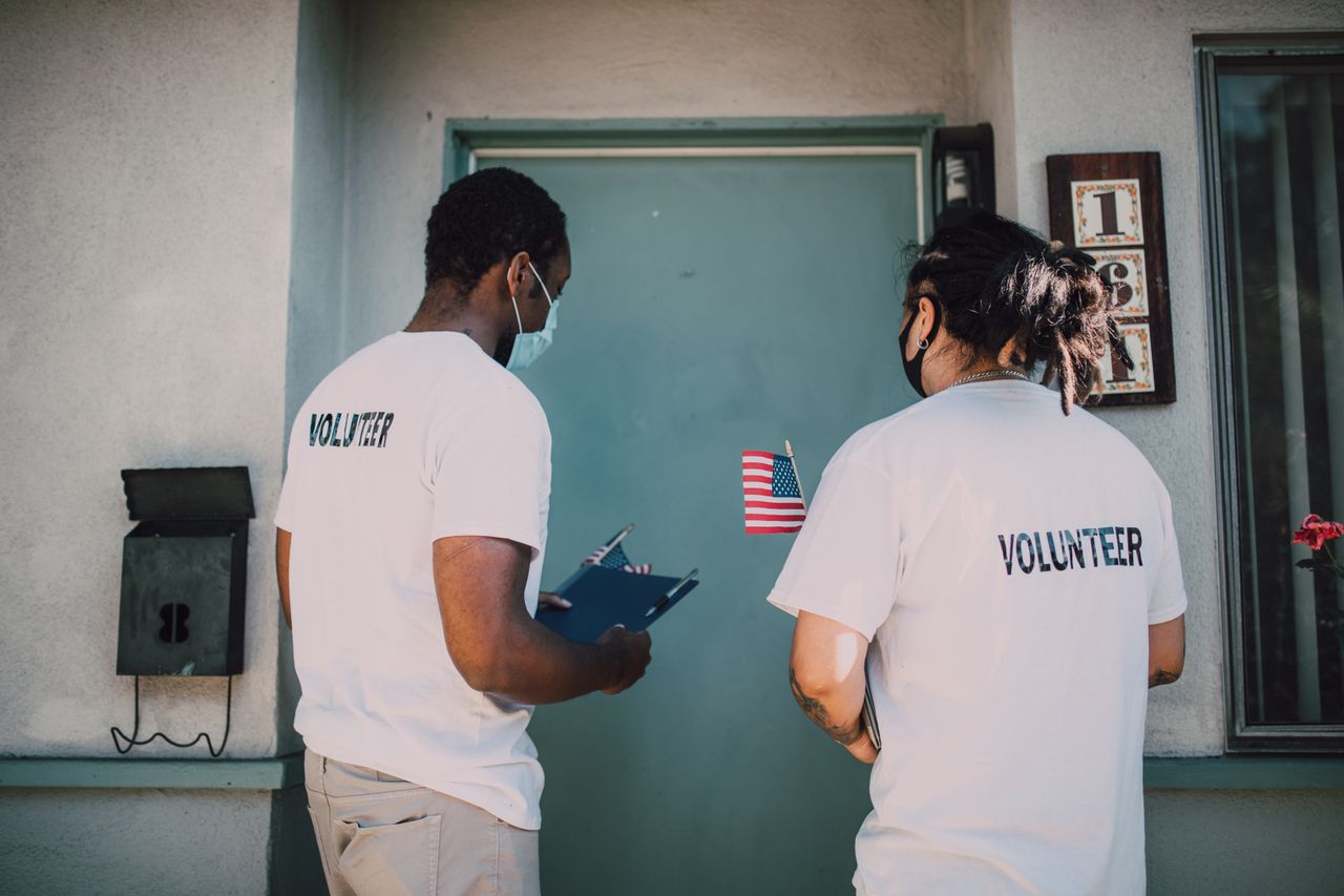 Two volunteers wear masks and visit community homes, engaging in outreach activities.